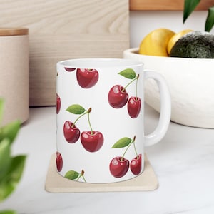 May include: White ceramic mug featuring a repeating pattern of ripe red cherries with green leaves. The mug has a curved handle and is set on a beige coaster. The background includes a wooden surface and a bowl of fruit.