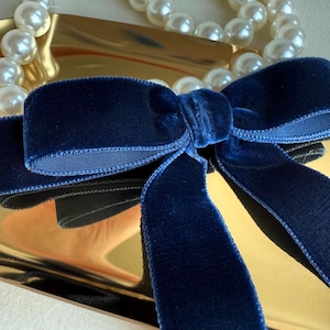 Puede incluir: Un bolso de mano de metal dorado con un lazo de terciopelo azul. El bolso tiene una correa de perlas.
