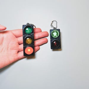 Może przedstawiać: Dwa miniaturowe breloki w kształcie sygnalizacji świetlnej. Większy brelok jest czerwony, żółty i zielony. Mniejszy brelok jest zielony, czerwony i ma symbol pieszego.