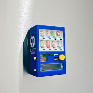 May include: A blue miniature vending machine with a yellow button and a clear display. The machine has various beverage options, with text in Japanese. The words "COFFEE SINCE 2022" are printed on the side. The machine is attached to a silver surface.
