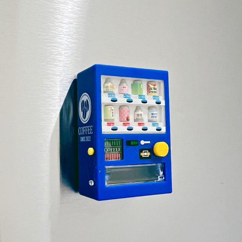 May include: A blue miniature vending machine with a yellow button and a clear display. The machine has various beverage options, with text in Japanese. The words "COFFEE SINCE 2022" are printed on the side. The machine is attached to a silver surface.