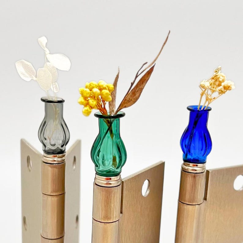 May include: Three decorative door hinges with small, colorful glass vases attached. The vases are gray, green, and blue, each holding dried flowers and foliage. The hinges are made of metal and are mounted on a white background.