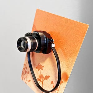 May include: A black camera with a silver lens and a black strap is mounted on an orange background. The word "Camera" is printed on the top of the camera. The background has a floral design.
