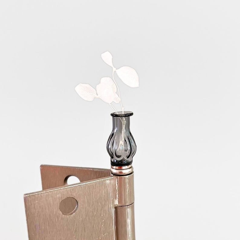 May include: A small, gray glass vase with a fluted design holds delicate white floral stems. The vase sits atop a silver-toned hinge, creating a unique decorative accent. The background is a soft, neutral white.