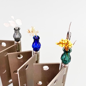 May include: A unique decorative display featuring three small glass vases in gray, blue, and green, each holding delicate dried flowers. The vases are mounted on metal hinges, creating an artistic arrangement against a white background.
