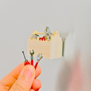 May include: Miniature wooden toolbox with assorted silver and red tools. The toolbox contains a wrench, hammer, and other tools. A hand holds a screwdriver, wrench, and pliers. The tools are small and detailed.