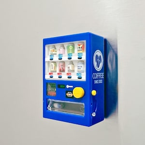 May include: A blue miniature vending machine with various beverage options. The machine has a yellow button and a coin slot. The text "COFFEE SINCE 2022" is visible on the side. The machine is mounted on a white surface.
