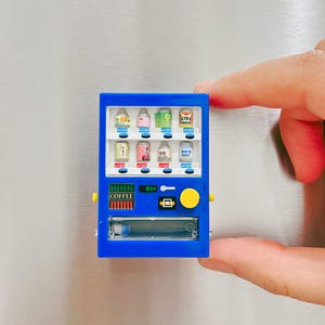 May include: A miniature blue vending machine with various beverage options, including canned drinks and bottled beverages. The machine has a "COFFEE" label and Japanese text. The vending machine is held by a person's fingers.