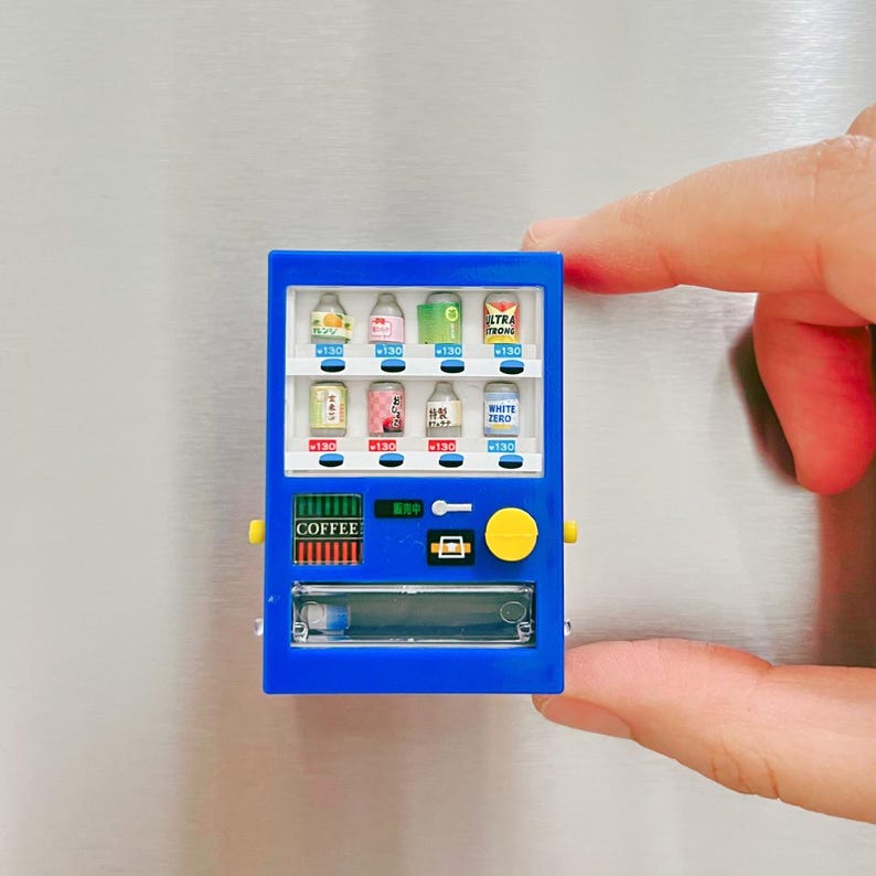 May include: A miniature blue vending machine with various beverage options, including canned drinks and bottled beverages. The machine has a "COFFEE" label and Japanese text. The vending machine is held by a person's fingers.