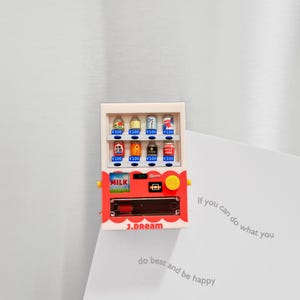May include: A red and white miniature vending machine with various canned drinks. The machine has a yellow button and the word "MILK" on the front. The text "J.DREAM" is printed on the bottom. The vending machine is a refrigerator magnet.