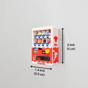 May include: A miniature white vending machine with red and blue accents, featuring various small product replicas. The machine is 2 inches (5 cm) tall and 1.4 inches (3.5 cm) wide, with the text "MILK" and "J.dream" visible.