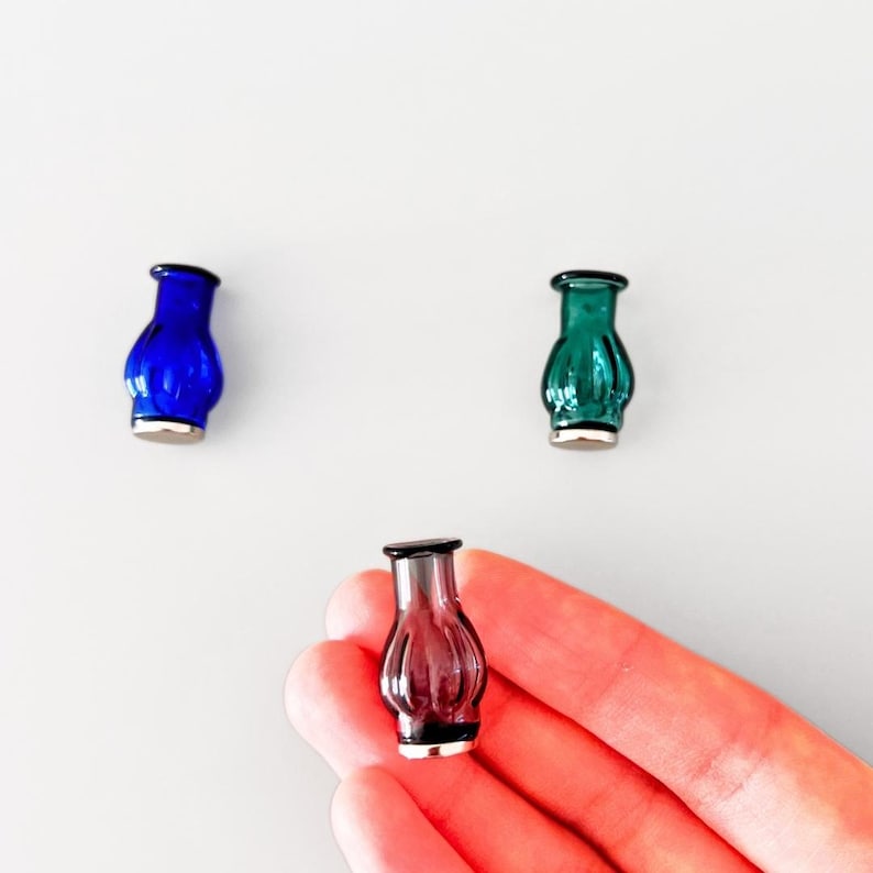 May include: Three miniature glass vases in blue, green, and purple. Each vase has a silver base and a dark rim. The vases are small, decorative items, likely for display or as part of a collection. The background is a plain white surface.