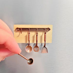 May include: A miniature kitchen utensil rack with five hanging utensils, including a ladle, a slotted spoon, a spatula, and two spatulas. The rack is made of wood and metal and has a rustic brown finish.