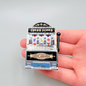 May include: A silver and black miniature slot machine with a lever. The machine has three spinning wheels with fruit symbols and the words "Lucky Slots" and "Lucky Jackpot".