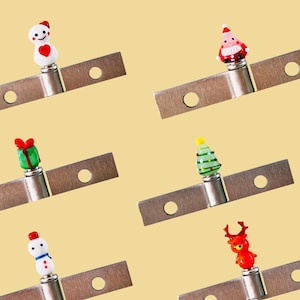 May include: A selection of Christmas bottle stoppers. Each stopper has a festive design, such as a snowman, Santa Claus, a gift, a Christmas tree, and a reindeer. The stoppers are attached to metal blades with a hole.