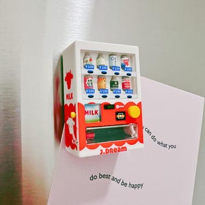 May include: A miniature white and red vending machine with various milk product options. The machine has a cow graphic and the text "MILK". The text "J.DREAM" is visible. A pink card with text is behind the machine.