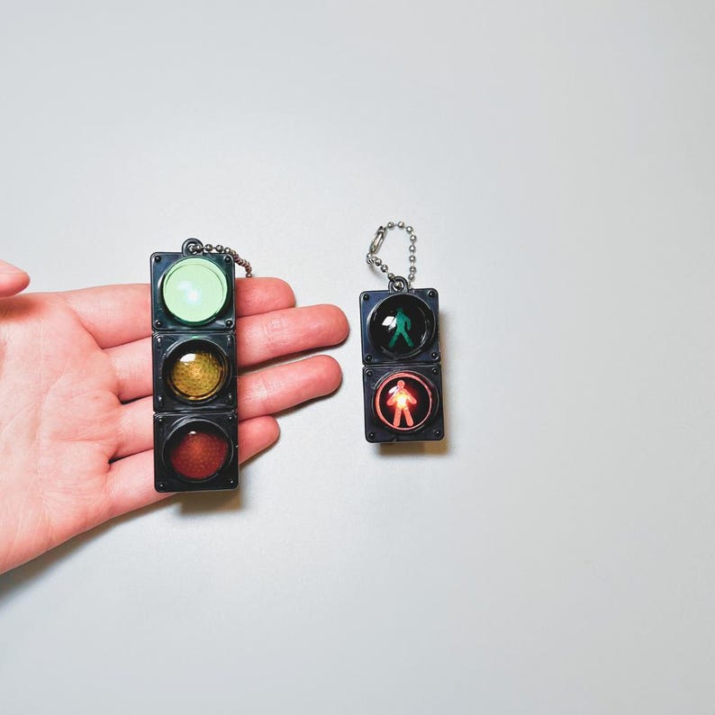 May include: Two traffic light keychains, one red, one green, with a walking person symbol. The keychains are black and have a silver chain.