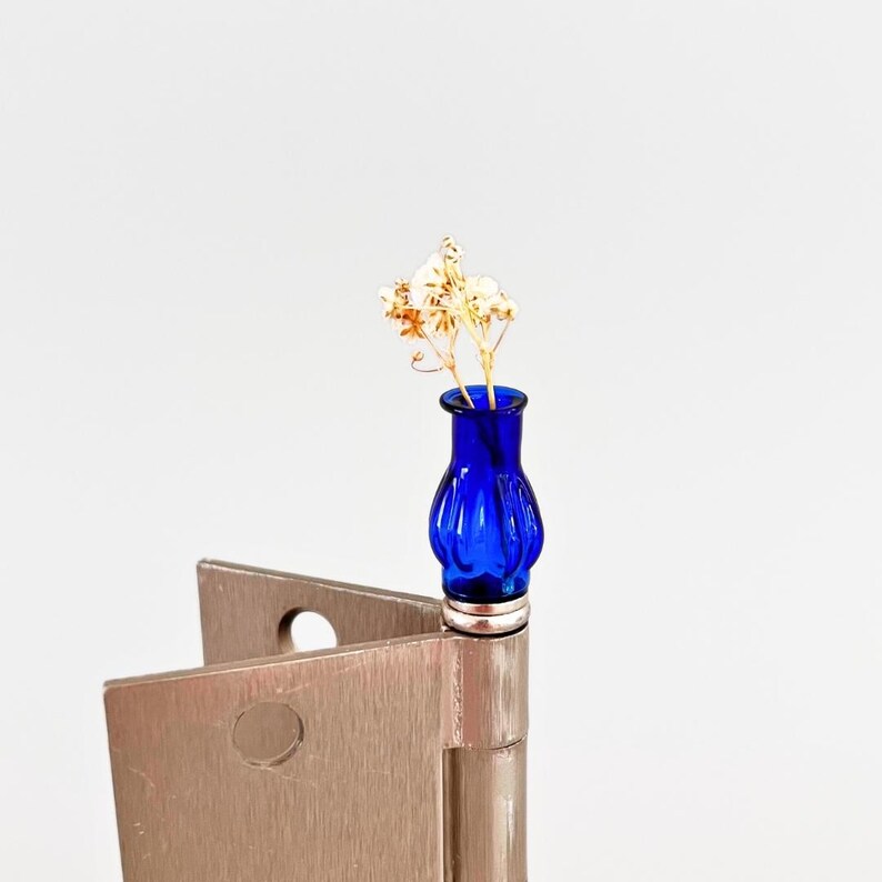 May include: A small, cobalt blue glass vase with a silver base, holding delicate dried flowers. The vase has a ribbed design and sits atop a metal hinge, creating a unique juxtaposition of textures and materials.