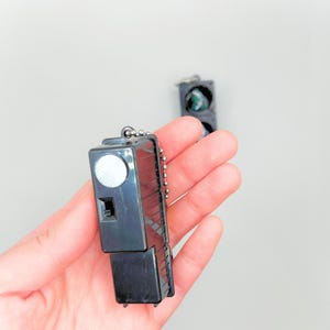May include: A black keychain shaped like a traffic light with a silver chain. The light is red, yellow, and green. The keychain is held in a hand.