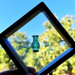 May include: A small, teal-colored glass vase with a black rim and base, framed within a black square. The vase is in focus against a blurred background of green foliage and a blue sky. The vase has a ribbed design.