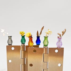 May include: A display of miniature glass vases in various colours, each containing a small flower or dried plant. The vases, in shades of grey, green, yellow, blue, and purple, are arranged on a brass hinge, creating a unique decorative piece.