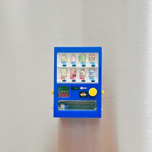 May include: A miniature blue vending machine with various colorful beverage cans displayed inside. The machine has a yellow button and the word "COFFEE" is visible. The background is a metallic surface.