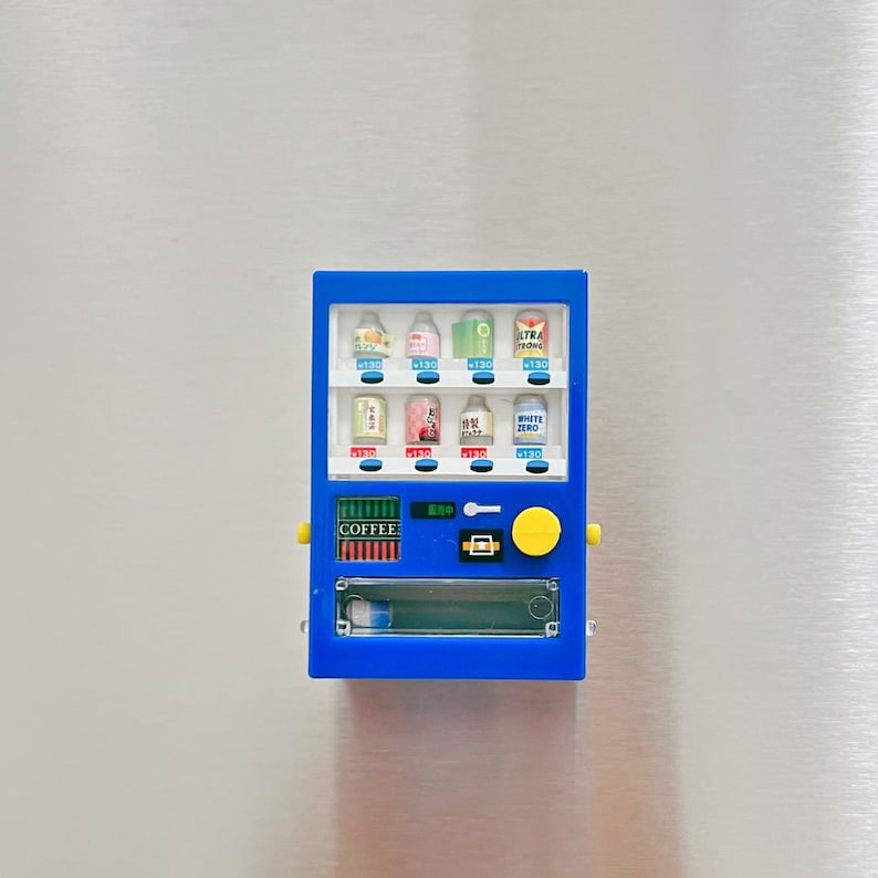 May include: A miniature blue vending machine with various colorful beverage cans displayed inside. The machine has a yellow button and the word "COFFEE" is visible. The background is a metallic surface.