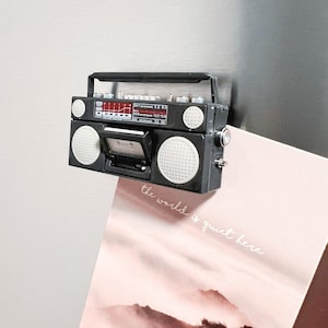 May include: A black miniature boombox magnet with white speaker covers and a red display. The cassette door is open, revealing the interior. The magnet is attached to a metallic surface, with a pink card that reads "the world is quiet here".