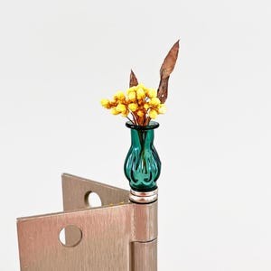 May include: A small, teal glass vase filled with yellow flowers and dried leaves. The vase sits atop a silver hinge, creating a unique and artistic display. The background is plain white.