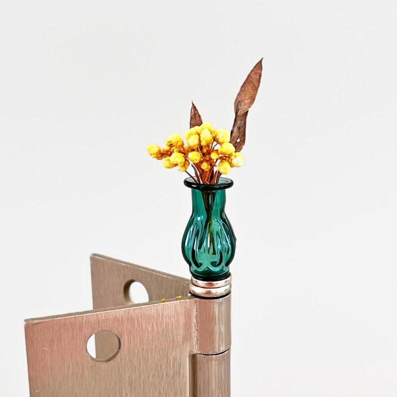 May include: A small, teal glass vase filled with yellow flowers and dried leaves. The vase sits atop a silver hinge, creating a unique and artistic display. The background is plain white.