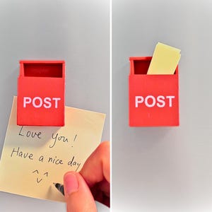 Puede incluir: Un buzón rojo en miniatura con la palabra "POST" en blanco. Una nota manuscrita dice "Love you! Have a nice day" con una carita sonriente. El buzón está sobre una superficie gris.