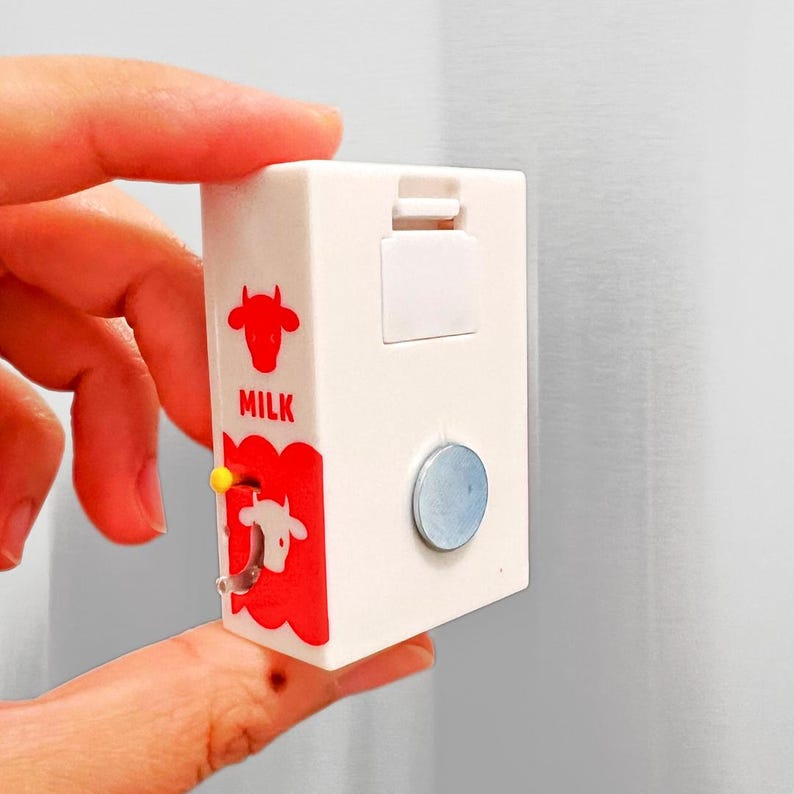 May include: A white milk carton-shaped magnet with red cow graphics and the word "MILK". The carton has a small, yellow, circular knob and a silver-colored magnet on the side.