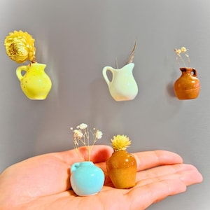 Könnte beinhalten: Fünf Miniatur-Keramikvasen in verschiedenen Farben und Stilen, darunter gelb, weiß, braun und blau. Einige Vasen sind mit Trockenblumen gefüllt.