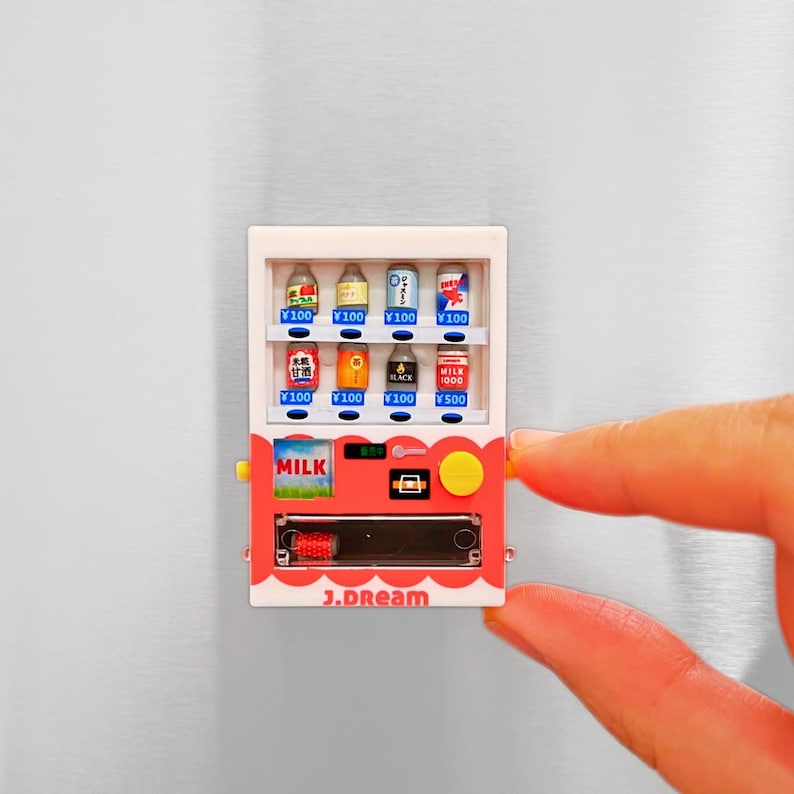 May include: A miniature vending machine with a red and white design, featuring various beverage options. The machine has a "MILK" label and displays prices in Japanese Yen. The text "J.DREAM" is printed at the bottom.