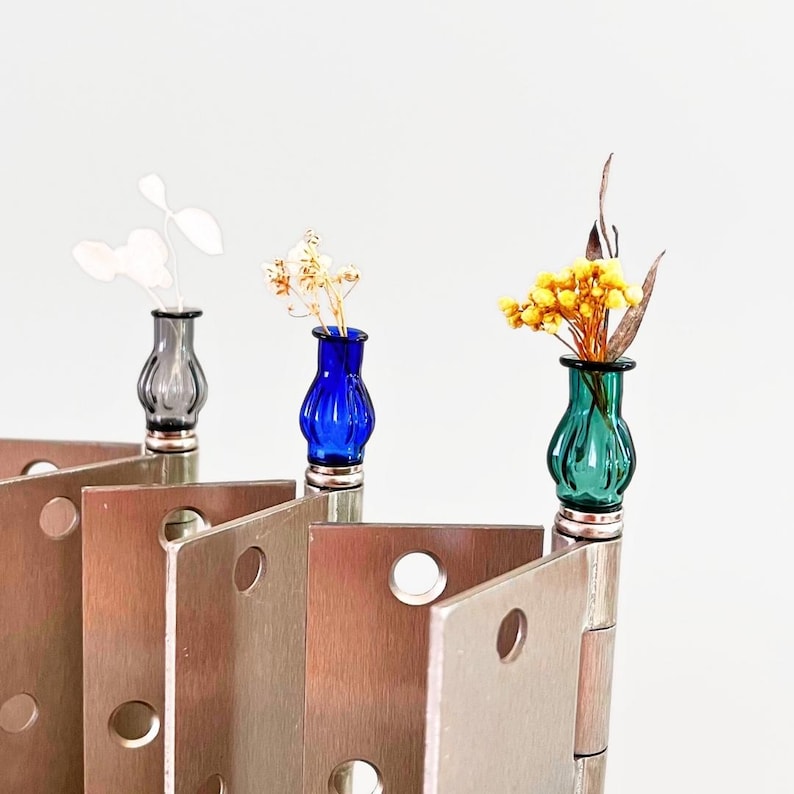 May include: Three small, colorful glass vases with dried flowers sit atop a series of metal hinges. The vases are blue, green, and gray, with silver bases. The hinges are a warm, metallic color, and the background is white.