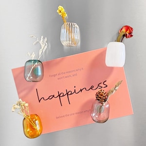 May include: A collection of small glass vases, each holding a dried flower or plant, arranged around a pink card with the word "happiness" and inspirational text. The vases are in various shapes and colors.