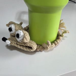 May include: A green tumbler rests on a handmade crochet coaster shaped like a whimsical creature. The coaster is beige with black and white accents, and the tumbler has a green handle.