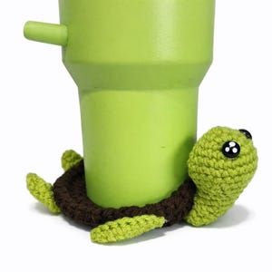 May include: A light green tumbler boot featuring a crocheted turtle design. The turtle has a dark brown shell, light green head, limbs, and black plastic eyes with white highlights. The accessory fits snugly around the base of a lime green tumbler.