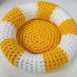 May include: A circular, bowl-shaped crochet item featuring a thick, padded rim with alternating yellow and white vertical stripes. The center base is a solid yellow, flat, circular crochet disc, creating a small, rounded, nest-like structure.
