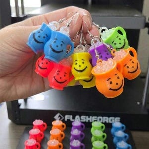 May include: A collection of colorful pitcher-shaped earrings featuring smiling faces and clear, textured liquid details. The earrings are available in blue, red, orange, yellow, purple, and green. Each piece includes a silver-toned metal hook.