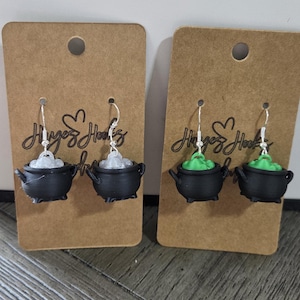 May include: Two pairs of dangle earrings featuring black cauldron charms on silver-toned hooks. One pair has white bubbling liquid, and the other has green bubbling liquid. Each pair is mounted on a rectangular brown card with the text HugsHooks.