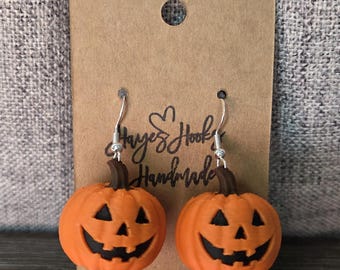 May include: A pair of orange pumpkin-shaped earrings with brown stems and black carved jack-o'-lantern faces. The earrings hang from silver-toned metal hooks and are attached to a rectangular brown cardboard display card labeled Hayes Hooks Handmade.