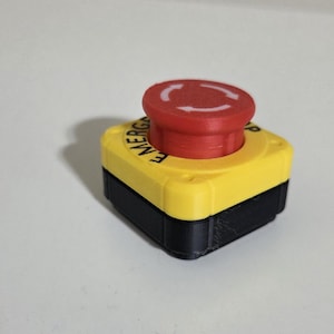 May include: A red emergency stop button with a yellow base and black housing. The button has a raised, circular top with a white arrow symbol. The word "EMERG" is visible on the yellow base.