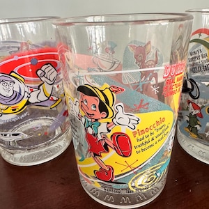 Op de afbeelding: Een doorzichtig glas met een kleurrijke afbeelding van Pinocchio en de tekst, Pinocchio had to be brave, truthful & unselfish to become a real boy. Het ontwerp bevat een gele zwaai, een klein hertje en de zin, Fantasy... lies beyond the stars.