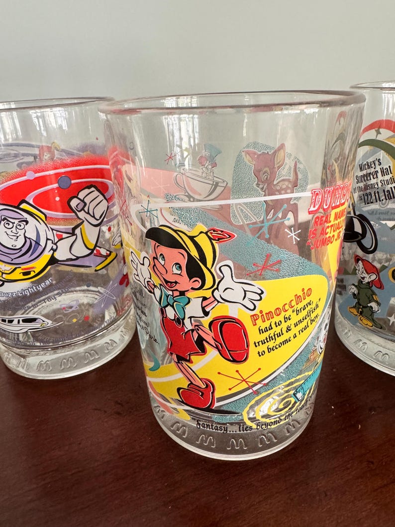 Op de afbeelding: Een doorzichtig glas met een kleurrijke afbeelding van Pinocchio en de tekst, Pinocchio had to be brave, truthful & unselfish to become a real boy. Het ontwerp bevat een gele zwaai, een klein hertje en de zin, Fantasy... lies beyond the stars.
