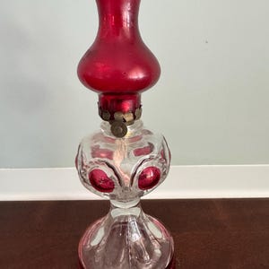 May include: A vintage oil lamp featuring a clear glass base with embossed circular patterns and red glass accents. The lamp includes a brass-colored metal burner assembly and a removable, ruffled-top red glass chimney globe for the flame.