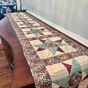 May include: A long, rectangular quilted table runner with a star pattern. The runner features cream-colored squares with teal and burgundy star designs, framed by a paisley border in burgundy, cream, and green. The runner is displayed on a dark wooden surface.