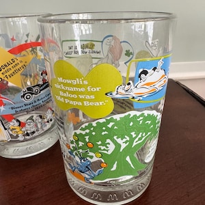 Op de afbeelding: Een helder drinkglas met kleurrijke Disney-karakterafbeeldingen en tekst. E&eacute;n afbeelding toont Mowgli en Baloe met de tekst, Mowgli's nickname for Baloo was Old Papa Bear. Een andere afbeelding toont een blauw insect dat met oranje ballen jongleert.