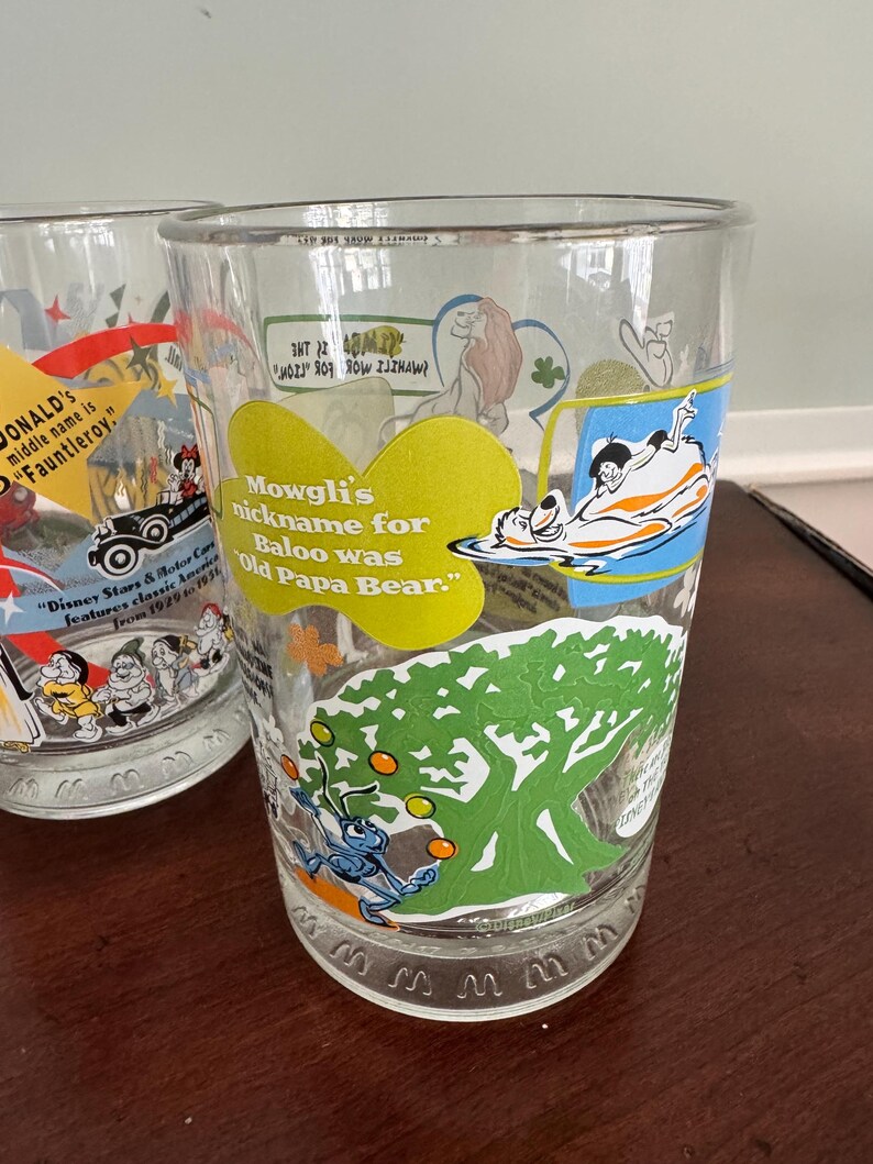 Op de afbeelding: Een helder drinkglas met kleurrijke Disney-karakterafbeeldingen en tekst. E&eacute;n afbeelding toont Mowgli en Baloe met de tekst, Mowgli's nickname for Baloo was Old Papa Bear. Een andere afbeelding toont een blauw insect dat met oranje ballen jongleert.