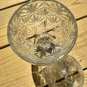May include: Clear glass serving bowl with a pedestal base and a matching lid. The bowl and lid feature a detailed, repeating starburst pattern. The bowl is round and sits on a short, hexagonal base. The lid is also round with a hexagonal handle.